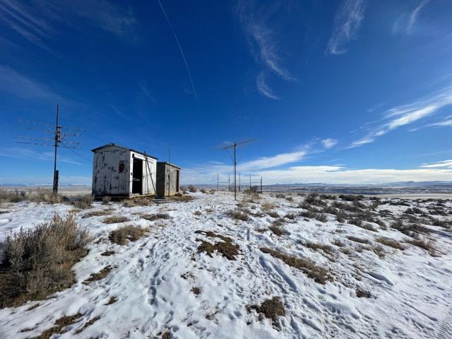 Two old radio tower buildings and two antennae are in an open area surrounded by snow on the ground and bright blue skies.