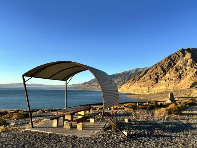 A covered picnic table on a concrete pad next to the shore of a blue lake with rocky hills.