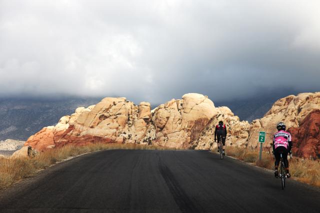 Two cyclists make their way up a smooth dark paved road backdropped by tan rock features.
