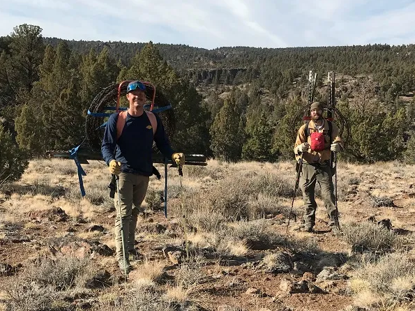 Two caucasian men pose outdoors in hiking clothes carrying metal stakes and wheels of wire on their backs.