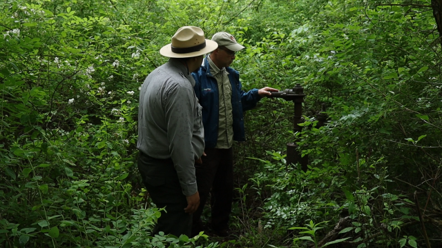 2 men stand next to a wellhead in a forested area, one rests his hand on it. 