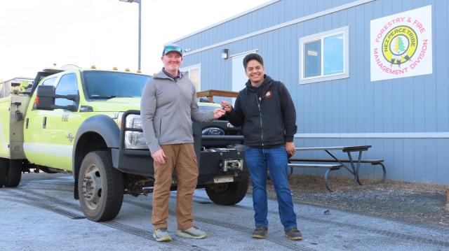 A young Caucasian man and a young Native American man smile while exchanging a set of keys in front of a yellow green truck