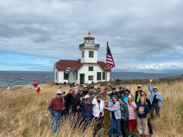 A large group of people are hoisting up an American flag and are standing in tall yellow grass with a while lighthouse in the background. They are on the edge of gray waters. 