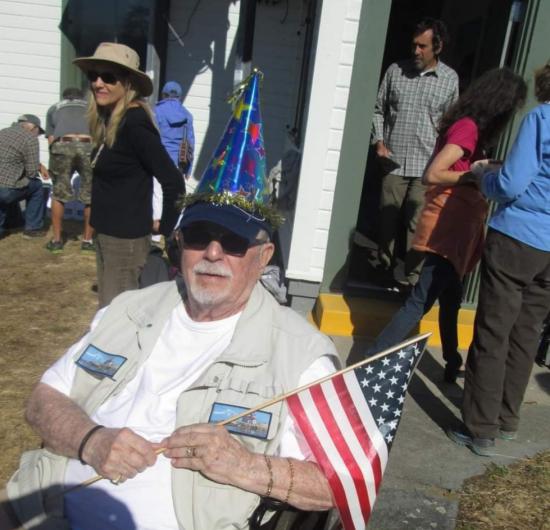 A man is sitting down holding a small plastic American flag and wearing a cone festive hat. People mill around in the background. 