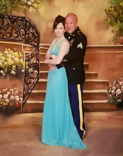 A Caucasian woman in a blue gown poses with a Caucasian man in military dress, embracing in front of a backdrop.