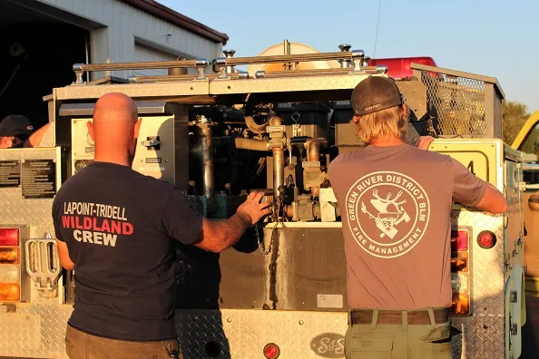 Two caucasian men, one in a tshirt reading Lapoint-Tridell Wildland Crew, the other in a tshirt reading Green River District BLM, work on the back of a fire engine.