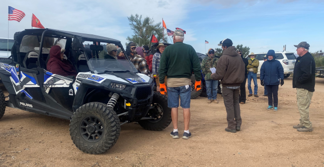 People standing in a semicircle receiving instruction for a desert cleanup with an off-road vehicle in the forefront.