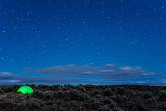 starry evening sky with green lit tent on sagebrush landscape