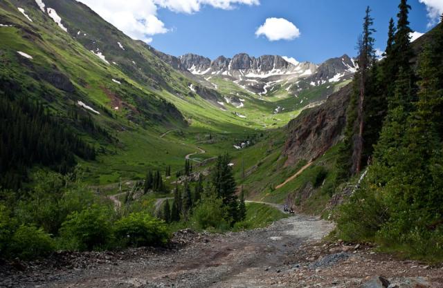 A trail weaves through a valley towards snowy mountains in the distance.