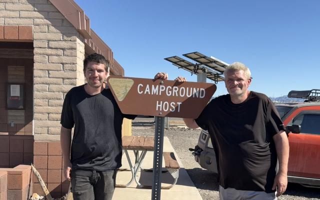Two men pose on either side of a sign contain the words CAMPGROUND HOST.