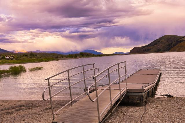 A photo showing a ramp with handrails leading to a fishing pier that juts out into the reservoir. The sky is cloudy and there are mountains in the background that are a mixture of shadow and light.