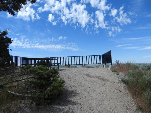 A photo showing a steep gravel trail leading to an overlook with a metal fence and a picnic table. Juniper trees are on the left side of the trail and native forbs on the right.