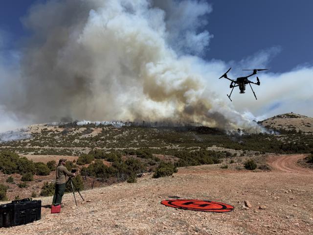 A large drove flies towards a fire while the pilot stands on the ground with the controls.