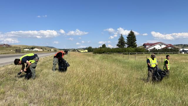 Photo of four men wearing yellow and orange safety vests and black rubber gloves picking up trash from the grassy shoulder of Highway 87.