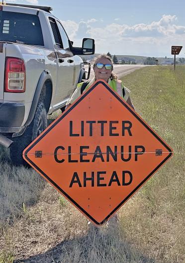 Photo of a smiling female BLM-NCMD employee holding a bright-orange sign reading "Litter Cleanup Ahead."