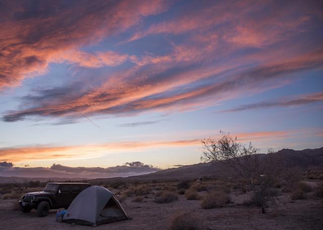 A jeep is parked next to a tent on a desert landscape, with a mountain in the background and the setting sun turning the sky and clouds pink and blue.