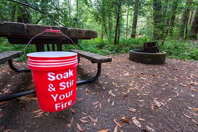 A bucket hanging from a picnic table and a fire pit in forested campsite.