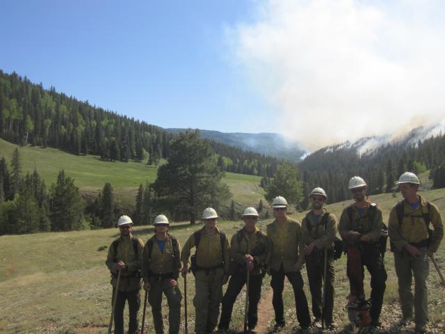 crew of 8 firefighters pose on a grass slope smoke is visible in the background