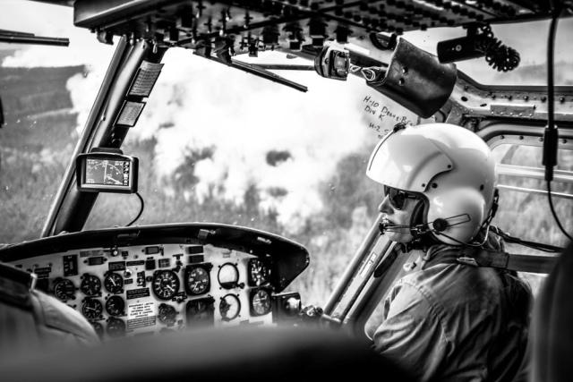 A black and white photo of a person sitting at the controls of a helicopter while flying over a landscape with smoke billowing up from a forest fire.