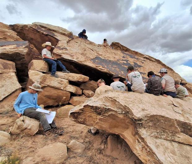 Young boys sit on boulders around a rock imagery site, grey clouds are overhead