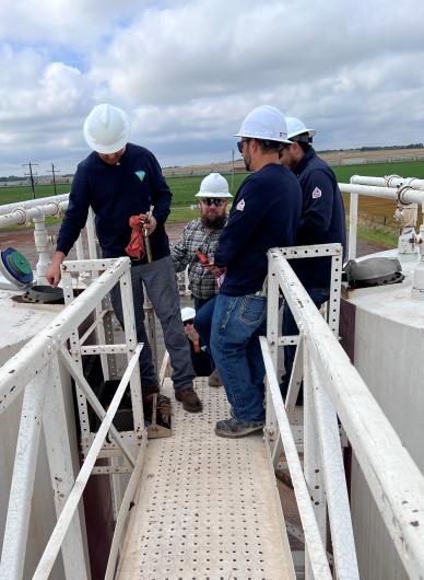 Four workers wearing white hard hats and navy shirts inspect equipment on an elevated metal platform outdoors with fields in the background.