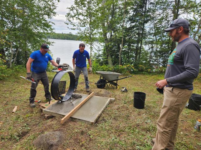 Three men work outdoors near a lake, constructing a concrete grill platform. Tools, buckets, and a wheelbarrow are visible.