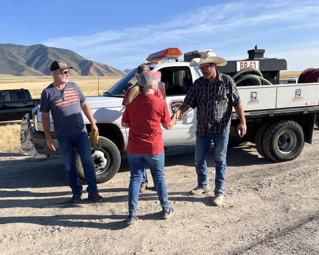 A woman shakes a man's hand in front of a truck on a dirt road with another man standing near by