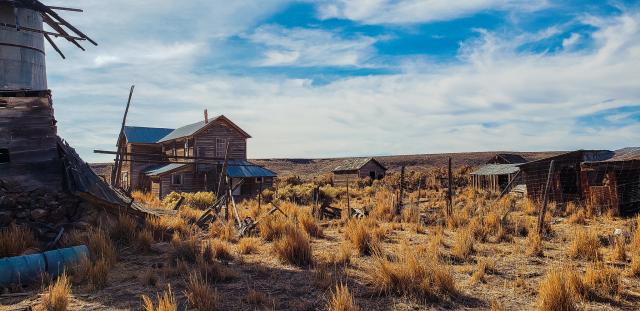 Abandoned wooden buildings and rusted structures scattered across a dry, grassy field under a partly cloudy blue sky.