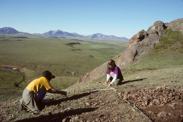 Two people working on a mountainside with a helicopter flying in the background