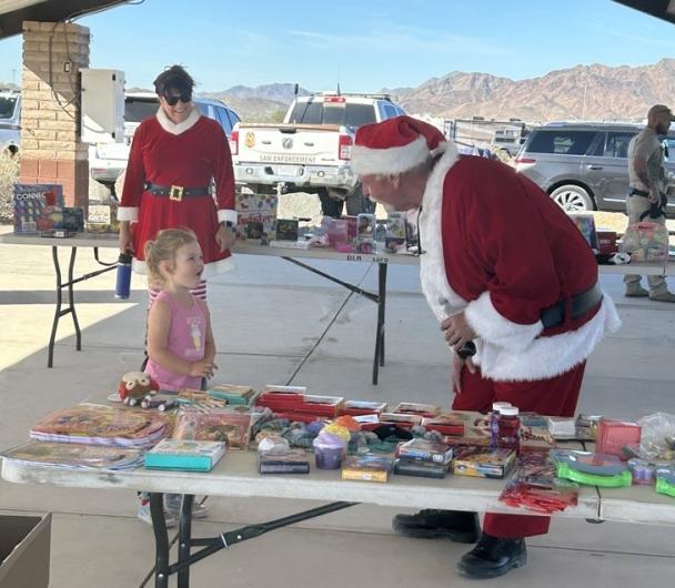 Mrs. Claus standing in the background watching a little girl visit with Santa Claus next to the table where children chose a present while visiting.