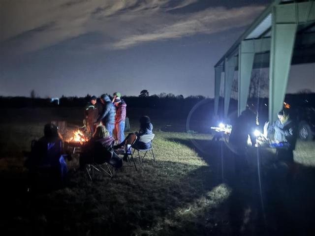 A group of people gathered around a campfire at night, with some sitting in chairs and others preparing food under a shelter.