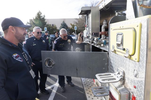 Men dressed in firefighting gear stand next to a fire engine, looking inside its opened compartments. 