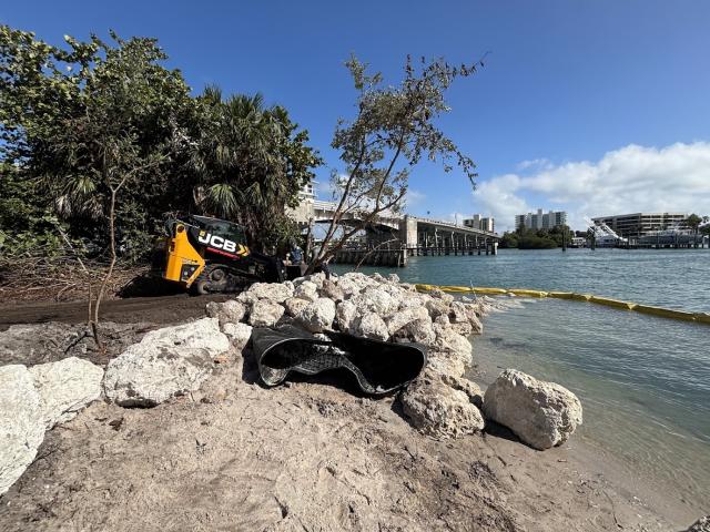 rocks piled along a sandy shoreline with construction equipment in the background