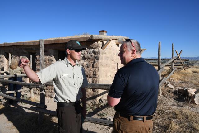 Two men talk in front of a stone house. 