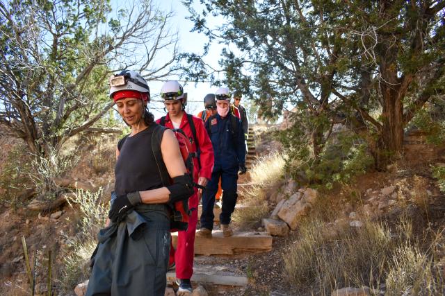 Six people wearing helmets and hiking gear walk down a set of steps in a wooded outdoor setting.