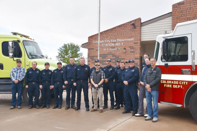 Firefighters in uniform stand in front of two large fire trucks one is green the other is red