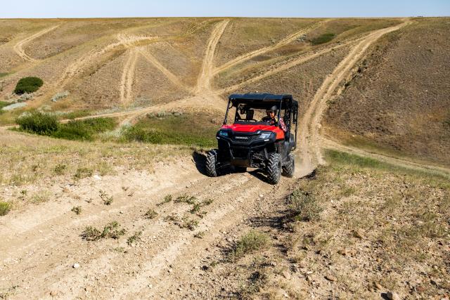 Red off-road utility vehicle driving on a dusty dirt trail through dry, hilly terrain under a clear sky.
