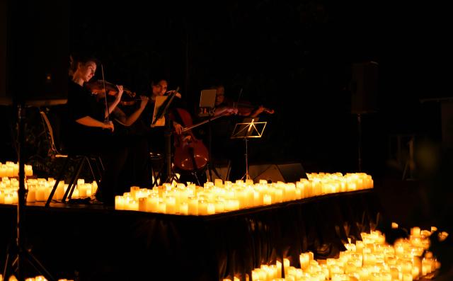 Musicians playing string instruments on a dark stage surrounded by numerous glowing candles, creating a warm, intimate atmosphere.
