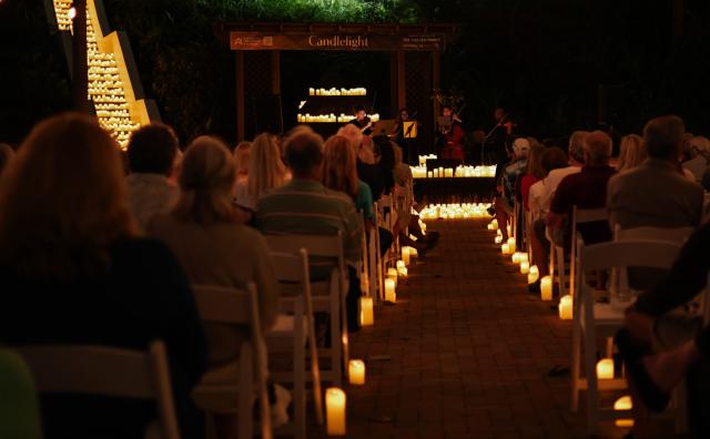 Audience seated on white chairs watches a string quartet performing at a candlelit outdoor event at night.