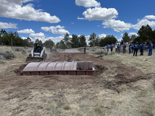 a group of people stand near a brown structure in the ground with machinery and dirt around it