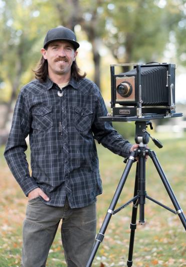 Man in flannel shirt poses for picture next to antique camera. 
