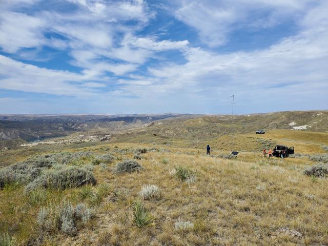 Wide view of a grassy, sagebrush-covered landscape beneath a partly cloudy blue sky. A slender Motus antenna tower stands on a distant ridge near several people, equipment and parked vehicles, with rugged river-breaks terrain stretching across the background.