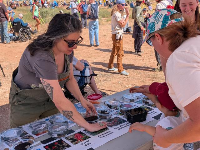 a woman speaks to a woman and a child. Several images of plants are on a table.