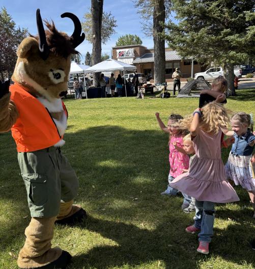 Children jump and raise their arms alongside Seymour the Antelope during Kemmerer Outdoor Discovery Day at Triangle Park in Kemmerer