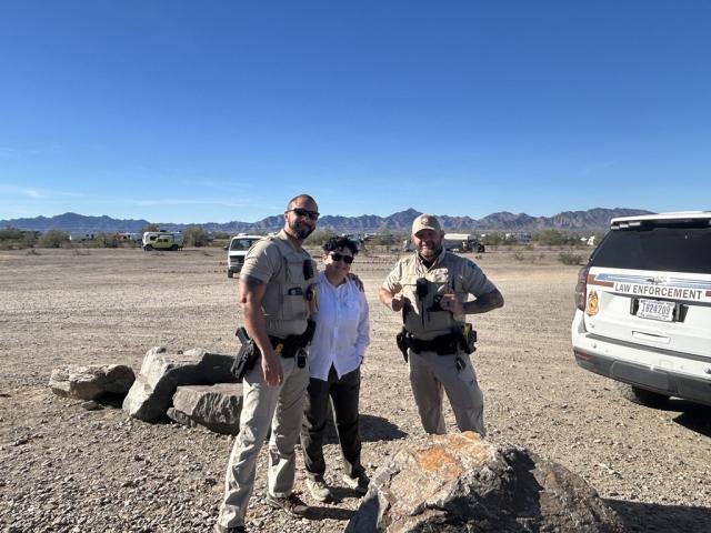 Two uniformed BLM rangers stand on either side of their civilian co-worker with a desert campground in the background. 