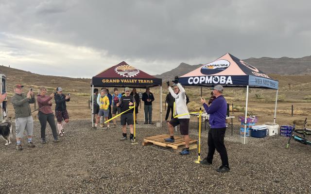 A couple dozen people stand around clapping while a person cuts a yellow ribbon; a cloudy desert landscape behind
