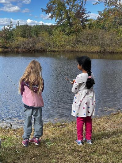 Two children stand along the edge of a pond holding fishing rods and casting lines into the water during an outdoor recreation event.