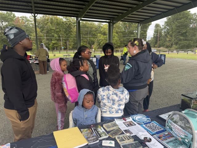 A Bureau of Land Management educator speaks with a group of children and adults gathered around a wildlife education table beneath an outdoor pavilion. Educational displays, insect specimens, and nature activity materials are spread across the table.