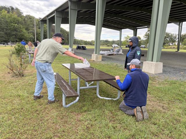Volunteers assemble a metal picnic table beneath a large open-air pavilion in a park setting with grassy fields and trees nearby.