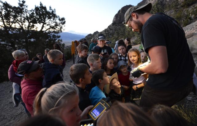 A group of children and adults gathers along a rocky mountain trail at dusk while a wildlife educator uses a flashlight to demonstrate or explain something held in gloved hands. The audience stands closely around the presenter, with several children watching attentively. Trees, rugged cliffs, and a broad desert valley form the background, and one child holds a handheld device displaying a colorful screen. The outdoor educational program takes place in fading evening light near a mountainous landscape.